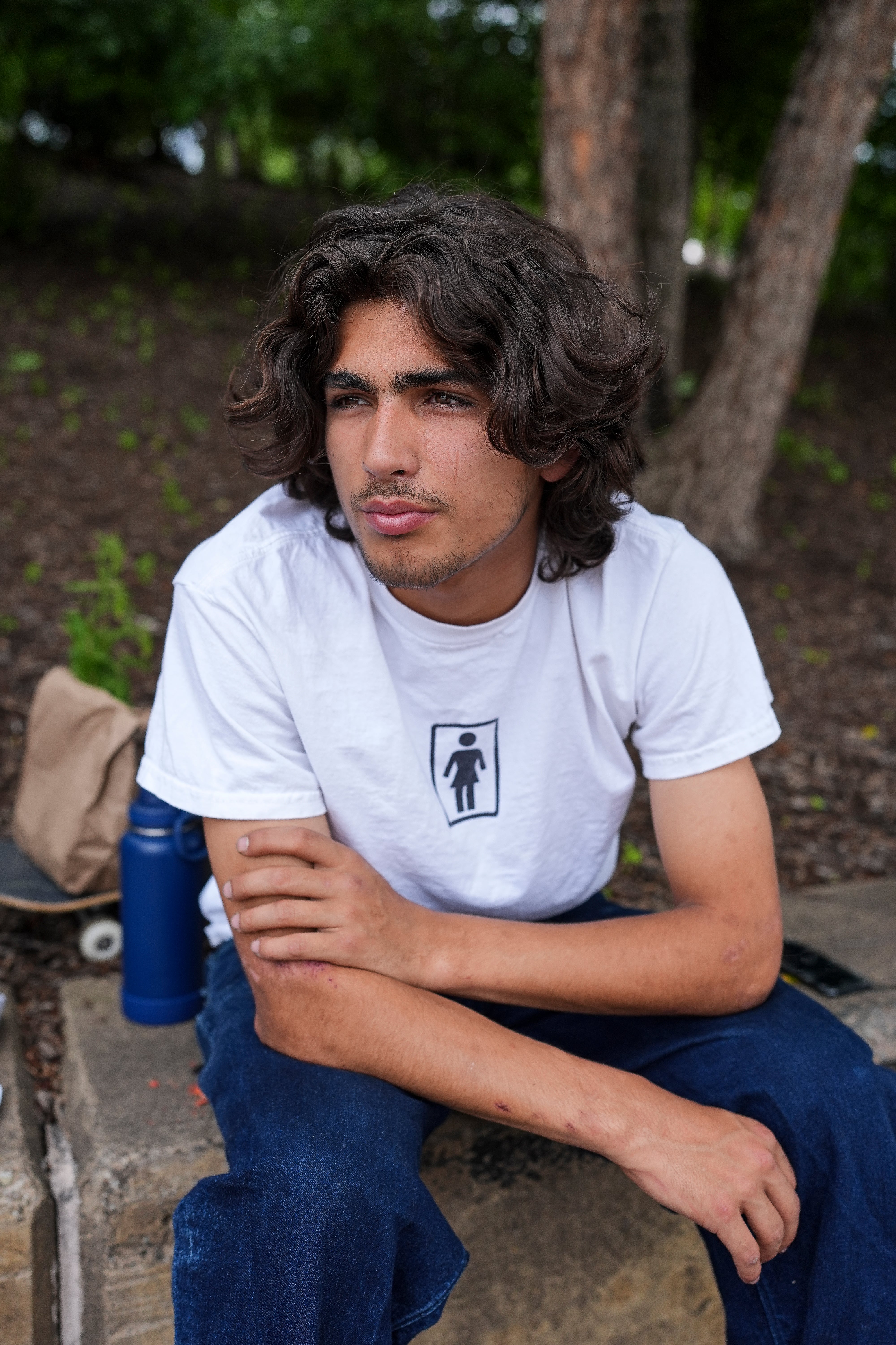 Person wearing a white t-shirt with a logo sitting outdoors near trees.