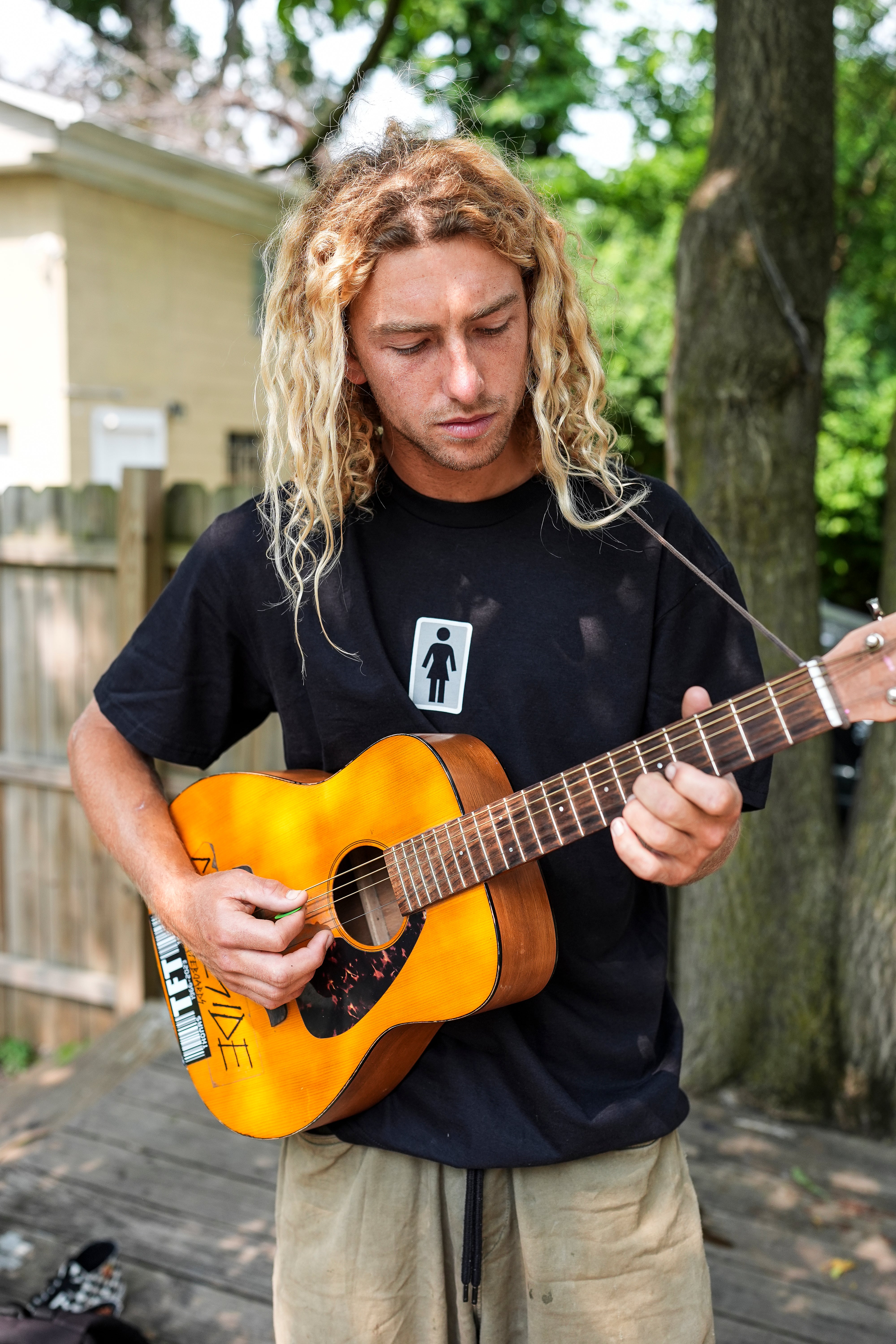 Person playing an acoustic guitar outdoors with trees and a wooden deck in the background