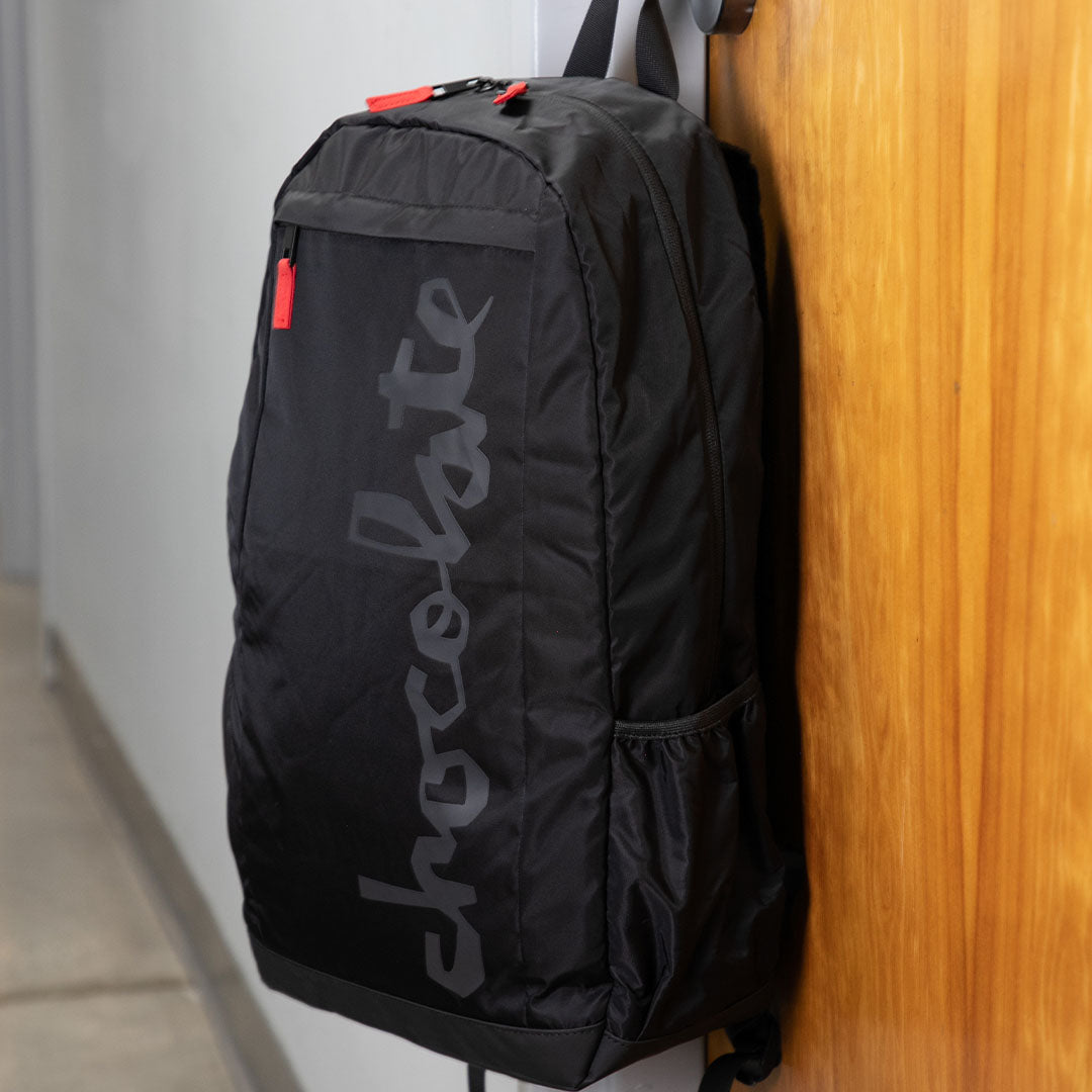 Black backpack with 'chocolate' branding against a wooden wall.